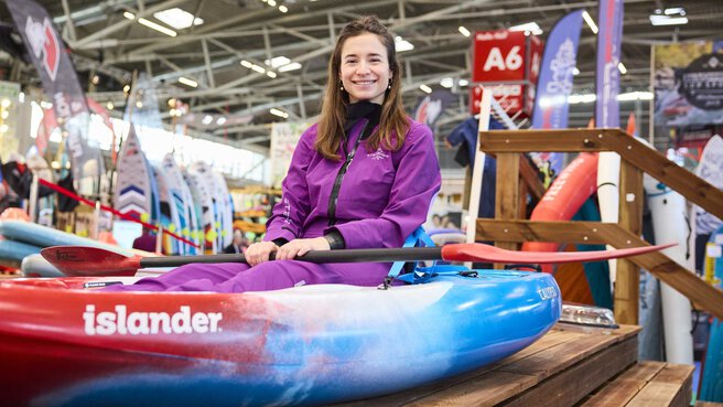 Lächelnde Frau im Kajak auf dem Indoor-Paddelbecken der Messe f.re.e, mit Paddel in der Hand. Es sind verschiedene Messestände im Hintergrund.