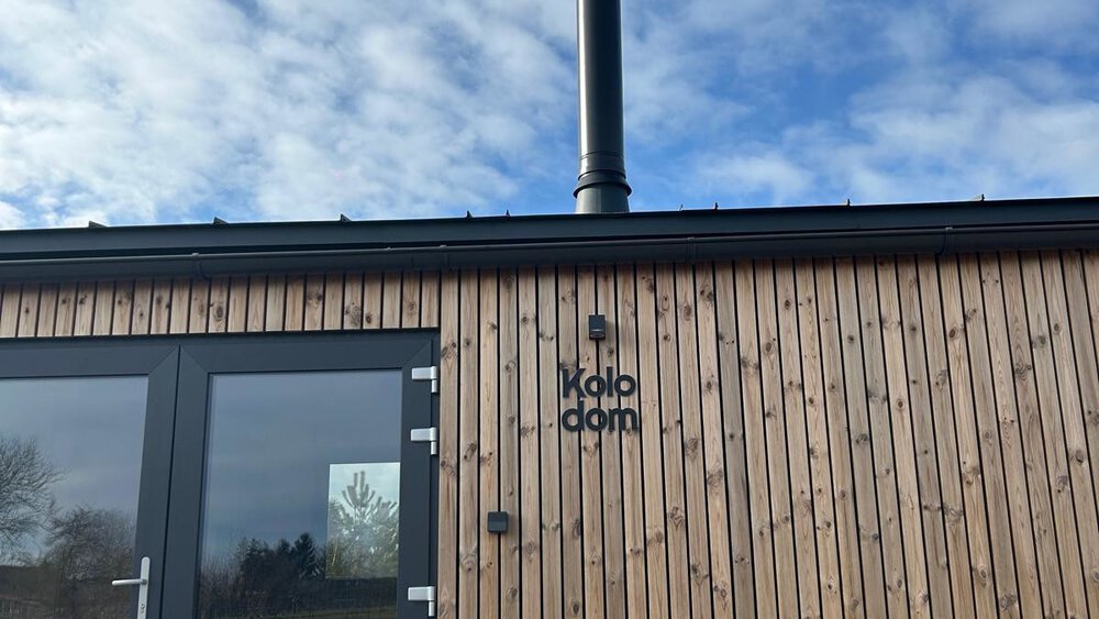 Kleine Holzhütte mit Kamin unter bewölktem Himmel. In der Glastür spiegeln sich Bäume und Landschaft wider.