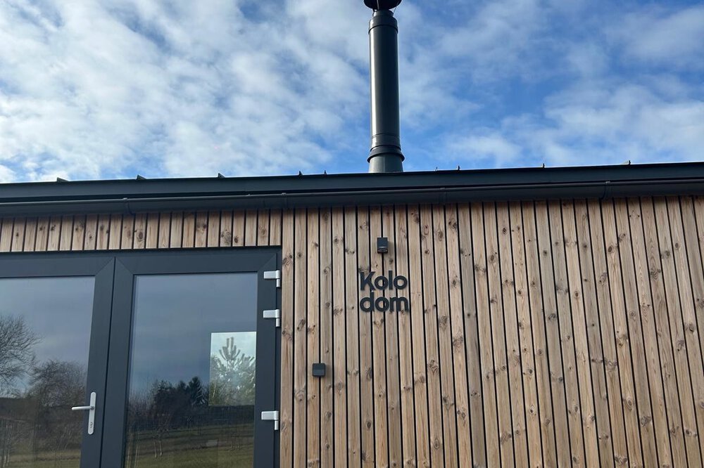 Kleine Holzhütte mit Kamin unter bewölktem Himmel. In der Glastür spiegeln sich Bäume und Landschaft wider.