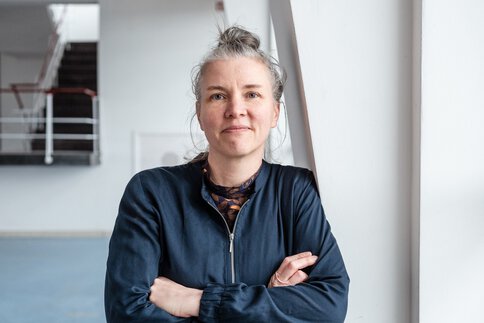 Frau mit grauem Haar in einem Dutt, trägt eine marineblaue Jacke, steht mit verschränkten Armen vor einer weißen Wand in einem Innenraum.