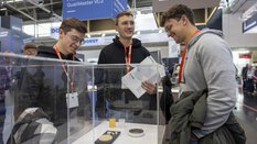 Drei Männer begutachten ein Exponat in einer Vitrine an einem Messestand. Sie sind leger gekleidet und halten Broschüren in den Händen.