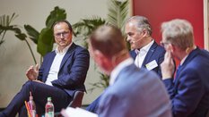 Vier Männer in Anzügen unterhalten sich bei einer Podiumsdiskussion. Ein Mann spricht gerade und ist im Fokus der Aufnahme, die Anderen sind leicht unscharf.