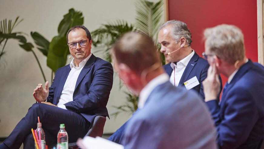 Vier Männer in Anzügen unterhalten sich bei einer Podiumsdiskussion. Ein Mann spricht gerade und ist im Fokus der Aufnahme, die Anderen sind leicht unscharf.