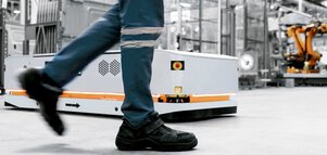 Person in work boots next to a small moving robot in an industrial setting.