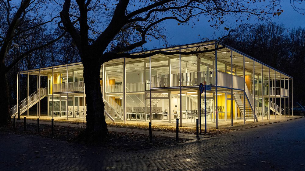 Modernes, zweistöckiges Glasgebäude, nachts beleuchtet, umgeben von Bäumen und einer gepflasterten Fläche.