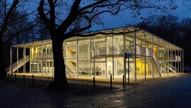 Modernes, zweistöckiges Glasgebäude, nachts beleuchtet, umgeben von Bäumen und einer gepflasterten Fläche.