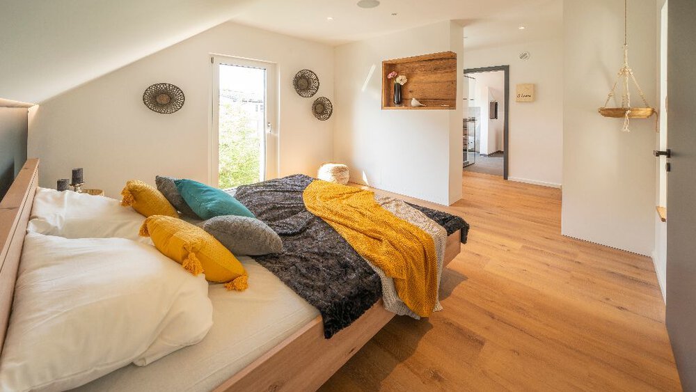 Gemütliches Dachgeschossschlafzimmer mit bunten Kissen, Dachschräge und Holzboden und Zugang zu einem kleinen Nebenzimmer.