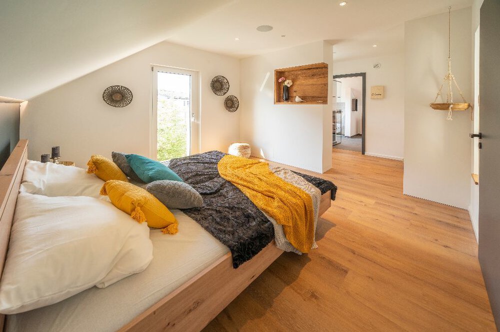 Gemütliches Dachgeschossschlafzimmer mit bunten Kissen, Dachschräge und Holzboden und Zugang zu einem kleinen Nebenzimmer.