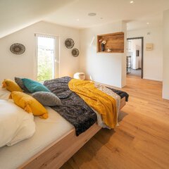 Ein Schlafzimmer mit einem großen Doppelbett aus Holz mit bunten Kissen, einem bodentiefen Fenster und einem schrägen Dach.