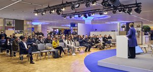 Expert talk on trade fair stage with female speaker at podium and audience in modern, well-lit conference area.