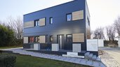 Modernes, kastenförmiges Haus mit dunkler Außenverkleidung, großen Fenstern und minimalistischem Design, umgeben von Bäumen.