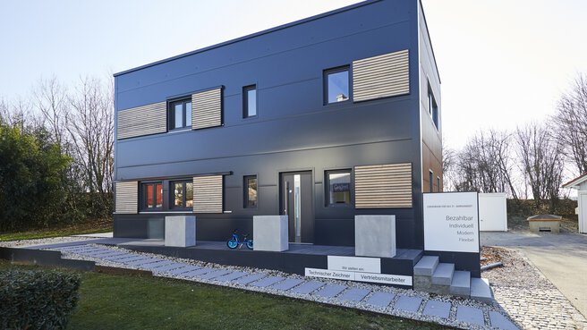 Modernes, kastenförmiges Haus mit dunkler Außenverkleidung, großen Fenstern und minimalistischem Design, umgeben von Bäumen.