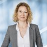 Eine lächelnde Frau mit gewelltem blondem Haar, in einem grauen Anzug und einem weißen Hemd.