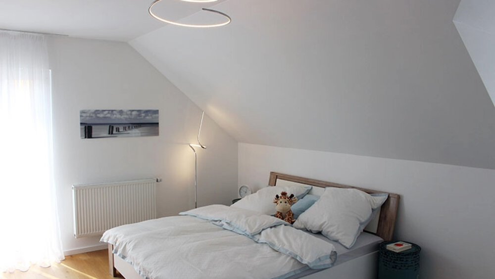 Schlafzimmer mit Dachschräge, weißen Wänden, einem Bett mit weißer Bettwäsche und moderner Beleuchtung.