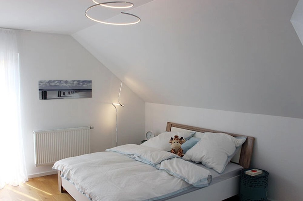 Schlafzimmer mit Dachschräge, weißen Wänden, einem Bett mit weißer Bettwäsche und moderner Beleuchtung.
