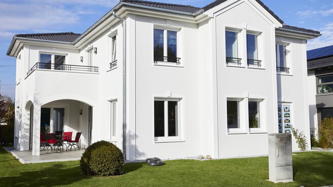Modernes zweistöckiges weißes Haus mit gepflegtem Rasen, gewölbter Terrasse und Balkon unter einem klaren blauen Himmel.