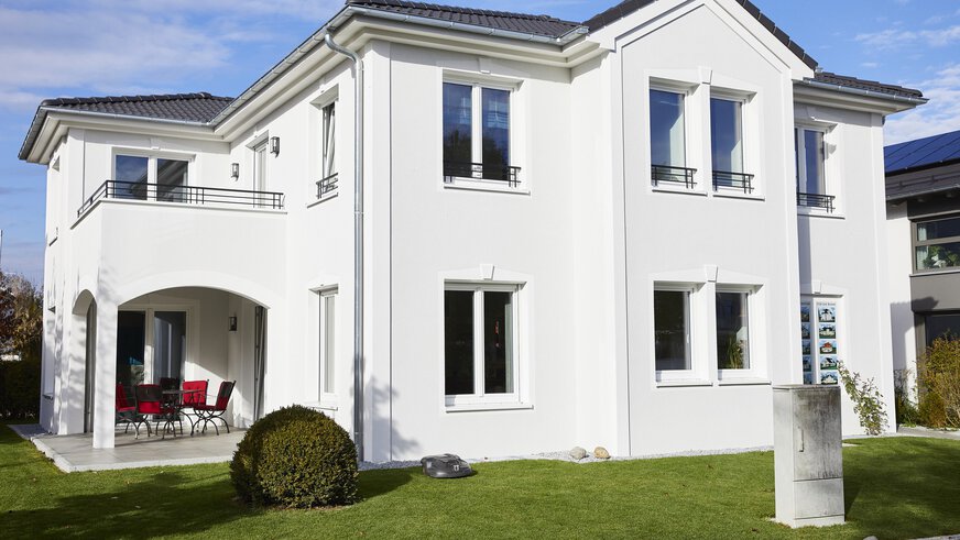 Modernes zweistöckiges weißes Haus mit gepflegtem Rasen, gewölbter Terrasse und Balkon unter einem klaren blauen Himmel.