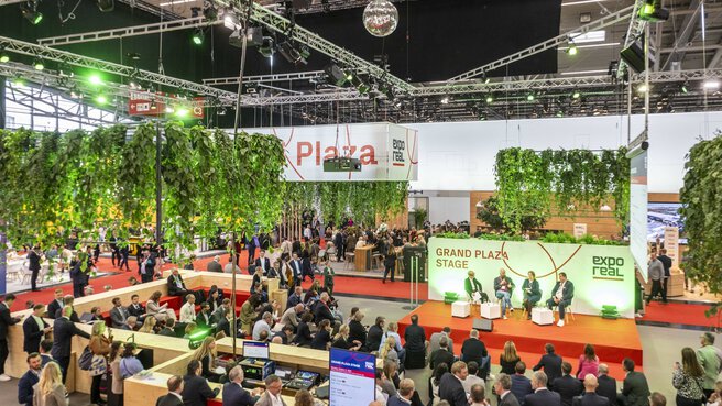 Blick auf die „Grand Plaza Stage“ der EXPO REAL in München, wo eine Podiumsdiskussion vor zahlreichem Publikum stattfindet. Die Bühne ist grün beleuchtet und von hängenden Pflanzen umgeben, die eine natürliche Atmosphäre schaffen. Im Hintergrund sind weitere Messestände, Sitzbereiche und Besucherinnen und Besucher zu sehen, die durch die Halle gehen oder Gespräche führen.