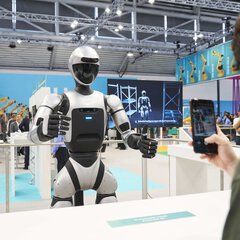 A humanoid robot stands at a trade fair as a person takes a photo with a smartphone.