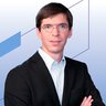 A man in a suit and glasses is standing with his arms crossed in front of a blue geometric background.