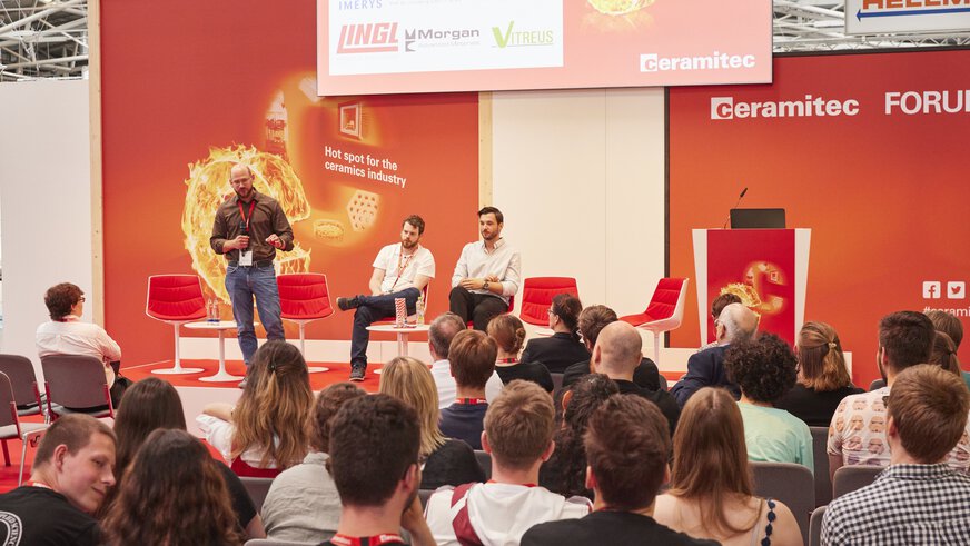 A speaker gesticulates in front of an audience on the stage of the ceramitec Forum, behind him sit two other speakers on red-postered chairs.