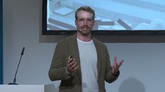 Eine Person steht am Rednerpult mit Mikrofon; Auf der Leinwand hinter ihr wird ein Bild gezeigt.