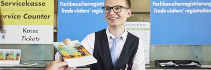 Smiling man with glasses behind the trade visitor registration counter, handing the observer an INTERFORST information brochure.