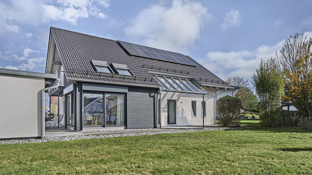 Das Musterhaus von KAMPA ist mit einem Satteldach und Photovoltaik-Anlage ausgestattet und wird durch große Fenster und einer mit Glas überdachten Terrasse ergänzt.