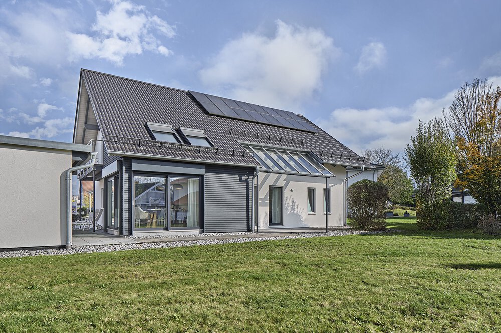 Das Musterhaus von KAMPA ist mit einem Satteldach und Photovoltaik-Anlage ausgestattet und wird durch große Fenster und einer mit Glas überdachten Terrasse ergänzt.