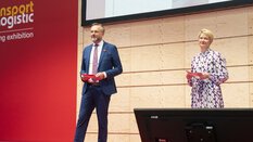 Ein Mann und eine Frau stehen auf einer Bühne der transport logistic und sprechen zum Publikum. Hinter ihnen hängt eine große Leinwand mit einer Liveaufnahme von der Bühne.