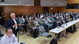 Ein Mann steht an einem Mikrofon im Publikum einer Veranstaltung der LOPEC und spricht zu jemand auf der Bühne, der nicht zu sehen ist.