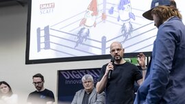 Fünf Personen auf einer Bühne, einer hält ein Mikrofon und spricht. Im Hintergrund ist eine Leinwand mit zwei boxenden Raketen in einem Boxring zu sehen.