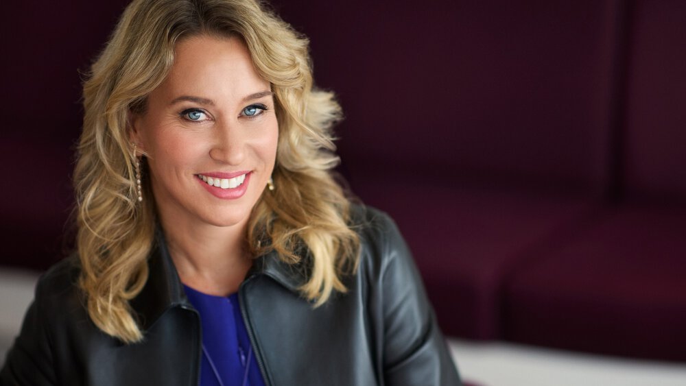 A smiling woman with blonde hair wearing a leather jacket, sitting on a purple couch.