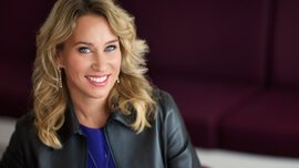 A smiling woman with blonde hair wearing a leather jacket, sitting on a purple couch.