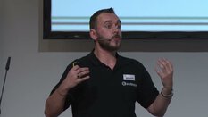 Ein bärtiger Mann im schwarzen Shirt spricht mit Headset und gestikuliert während einer Präsentation.