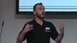 A bearded man in a black shirt speaks with a headset and gesticulates during a presentation.