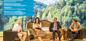 Eine Talkrunde zwischen einer Frau und drei Männern auf einer Bühne. Links oben im Bild ist ein blaues Schild mit Namen und Funktion der Anwesenden.