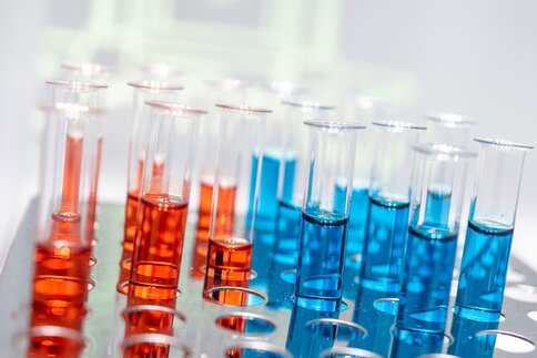 Multiple test tubes in a metal rack, filled with red and blue liquids, in a laboratory setting.