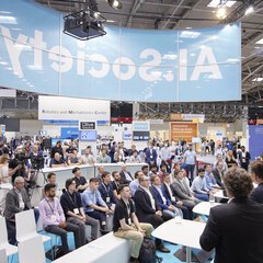 A large group of people sit in an exhibition hall in front of a stage with four speakers. Above them is a banner with the text 