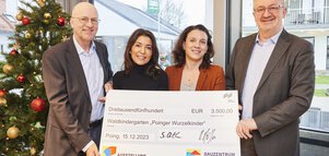 Vier Personen stehen vor einem Weihnachtsbaum in einem Haus und halten einen großen Scheck über 3,500€ für den Waldkindergarten 