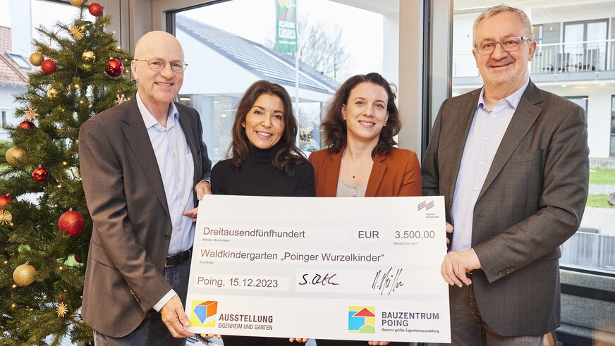 Vier Personen stehen vor einem Weihnachtsbaum in einem Haus und halten einen großen Scheck über 3,500€ für den Waldkindergarten 