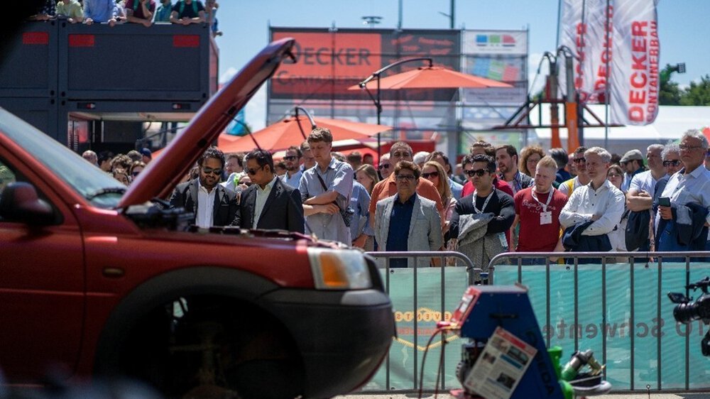 A group of people stands behind a barrier watching a live demonstration outdoors. In the foreground, the hood of a red car is open, with technical equipment next to it. Banners, parasols, and exhibition stands are visible in the background.