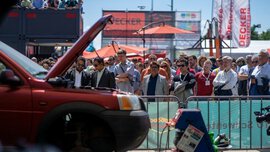 A group of people stands behind a barrier watching a live demonstration outdoors. In the foreground, the hood of a red car is open, with technical equipment next to it. Banners, parasols, and exhibition stands are visible in the background.