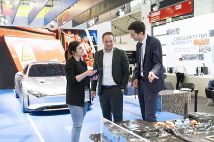 Two exhibitors stand in front of a car at a trade fair stand and demonstrate an engine block on display to a visitor.