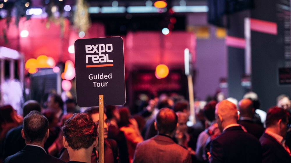 EXPO REAL 2026 in Munich: Numerous visitors and a guided tour, marked by an “EXPO REAL Guided Tour” sign, amid a lively trade fair atmosphere.