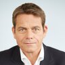 Eine lächelnder Mann im Anzug, mit kurzen braunen Haaren und intensivem Blick.