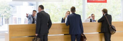 Menschen in Geschäftskleidung stehen am Empfangs- und Informationsschalter der EXPO REAL und sprechen mit dem Personal.