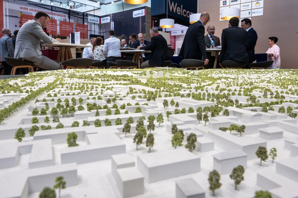 Großes Modell einer Stadt mit Bäumen und weißen Gebäudemodellen. Im Hintergrund sitzen Menschen in Geschäftskleidung an Tischen zusammen.