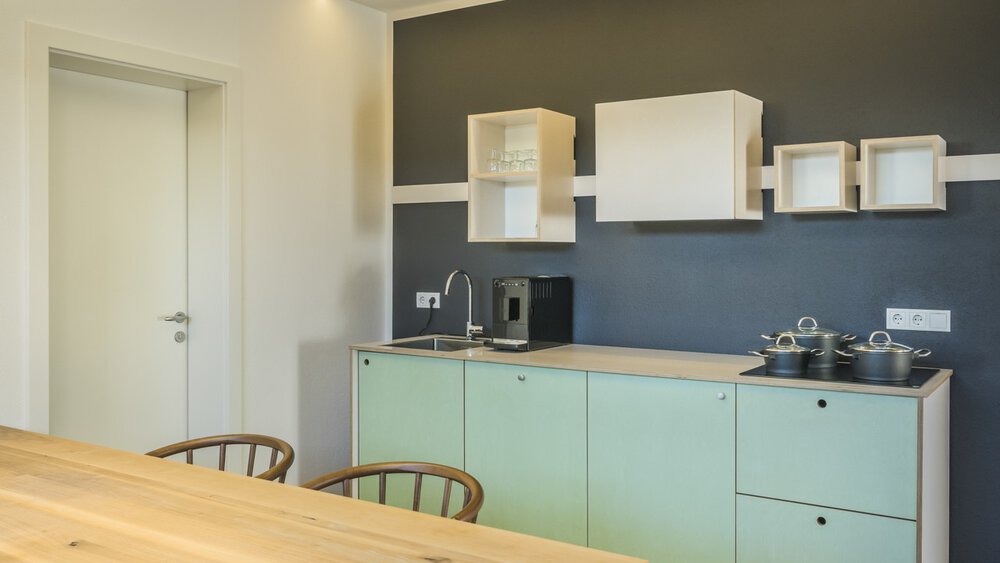 Moderne Küche mit hellgrünen Schränken, Esstisch aus Holz, dunkler Akzentwand und minimalistischem Dekor.