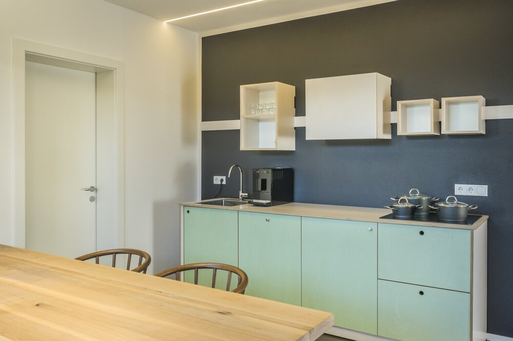 Moderne Küche mit hellgrünen Schränken, Esstisch aus Holz, dunkler Akzentwand und minimalistischem Dekor.